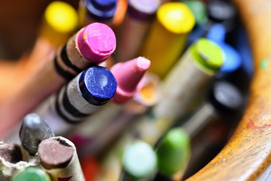 A close-up view of colorful crayons arranged in a container. The focus is on the tips of the crayons, showcasing a variety of colors including pink, blue, and yellow. The background is softly blurred, highlighting the vibrant colors of the crayons.