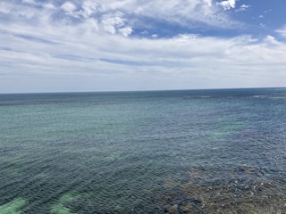 A vast expanse of ocean stretches towards the horizon, meeting a partly cloudy sky. The water is a mix of varying blue and green hues, with some visible seaweed beneath the surface.