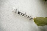 A textured white wall features the word 'threeology' written in a sleek, modern font with a transparent backing. A large green leaf is positioned to the right, adding a natural element to the minimalistic design.