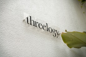 A textured white wall features the word 'threeology' written in a sleek, modern font with a transparent backing. A large green leaf is positioned to the right, adding a natural element to the minimalistic design.