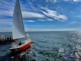 A sailboat with a red hull is afloat on a vast body of water under a bright blue sky. The sails are hoisted, and a person is seated at the rear of the boat. The sea appears slightly choppy with small waves reflecting the sunlight. A distant shoreline and scattered clouds are visible in the background.