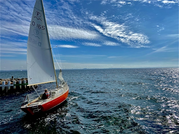 A sailboat with a red hull is afloat on a vast body of water under a bright blue sky. The sails are hoisted, and a person is seated at the rear of the boat. The sea appears slightly choppy with small waves reflecting the sunlight. A distant shoreline and scattered clouds are visible in the background.