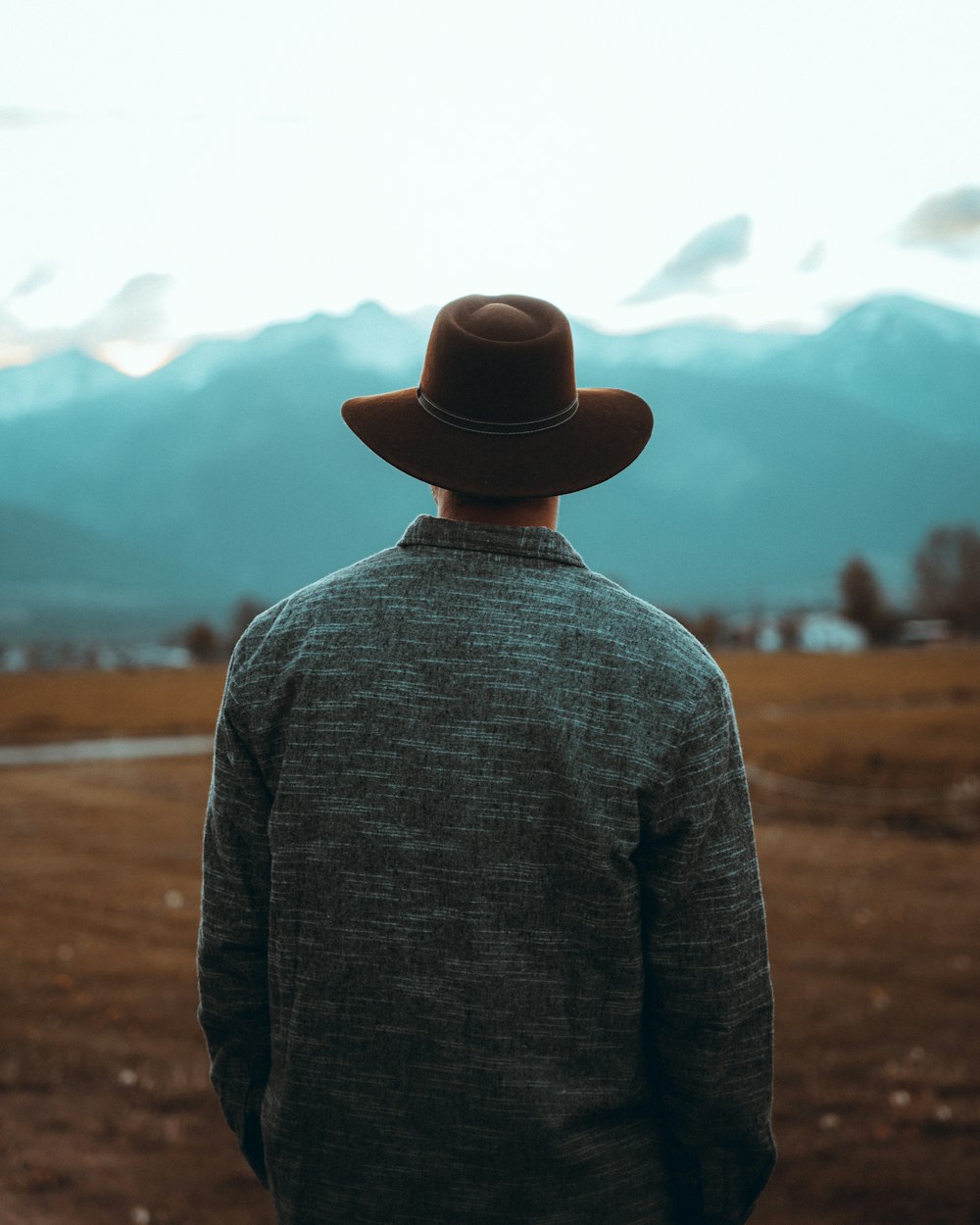 man in gray sweater and black fedora hat standing on brown field during daytime