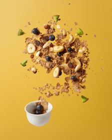 A dynamic composition of cereal, nuts, blueberries, banana slices, and avocado chunks appears to be suspended in mid-air above a small white bowl. The vibrant yellow background accentuates the colorful ingredients, creating a visually captivating scene.