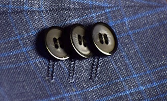 Three black buttons are sewn onto a dark blue fabric with a subtle plaid pattern featuring lighter blue lines. The buttons are aligned vertically and securely stitched, highlighting the texture and detail of the material.