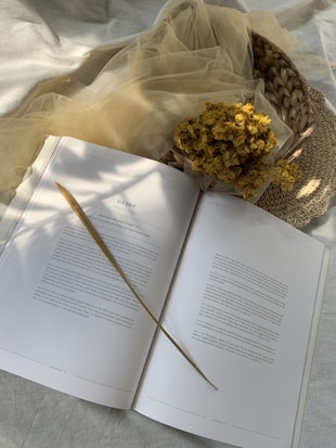 An open book with text is lying on a soft, light surface. A single long, dried grass stem rests across the pages, casting a subtle shadow. Beside the book, a cluster of dried yellow flowers sits on a woven basket adorned with beige, translucent fabric.