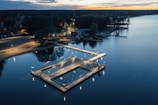 An aerial view of a serene outdoor swimming area located on a calm lake. The swimming section is illuminated with soft lights and is surrounded by a wooden deck. Nearby, a sandy beach and lush green forest can be seen under a twilight sky.