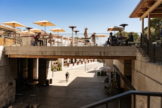 A modern outdoor terrace is bustling with people sitting at tables under large beige umbrellas. The terrace is situated on a concrete bridge-like structure, overlooking a spacious, open area below. The foreground includes a walkway leading down into the lower level. The scene is sunny, casting shadows and giving a clear view of the architecture.