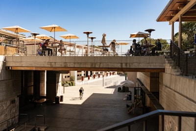 A modern outdoor terrace is bustling with people sitting at tables under large beige umbrellas. The terrace is situated on a concrete bridge-like structure, overlooking a spacious, open area below. The foreground includes a walkway leading down into the lower level. The scene is sunny, casting shadows and giving a clear view of the architecture.