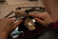 Hands holding tools are working on a metal piece with a chain and heart-shaped pendant inscribed with 'Hug me' on a workbench.