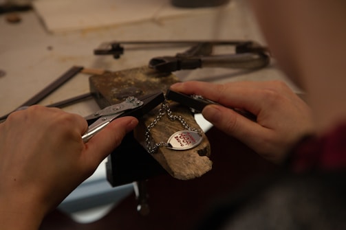 Hands holding tools are working on a metal piece with a chain and heart-shaped pendant inscribed with 'Hug me' on a workbench.