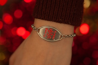 A close-up of a wrist adorned with a medical alert bracelet. The bracelet is chain-linked with a prominent metallic plate featuring a red medical symbol and the words 'Medic Alert' engraved on it. The background is composed of blurred, warm-toned lights, enhancing the focus on the bracelet.