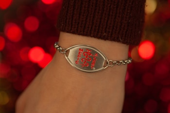 A close-up of a wrist adorned with a medical alert bracelet. The bracelet is chain-linked with a prominent metallic plate featuring a red medical symbol and the words 'Medic Alert' engraved on it. The background is composed of blurred, warm-toned lights, enhancing the focus on the bracelet.