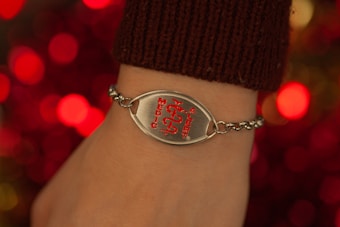 A close-up of a wrist adorned with a medical alert bracelet. The bracelet is chain-linked with a prominent metallic plate featuring a red medical symbol and the words 'Medic Alert' engraved on it. The background is composed of blurred, warm-toned lights, enhancing the focus on the bracelet.