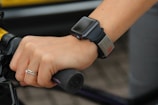 A hand gripping a bicycle handlebar is adorned with a smartwatch and a wristband. The smartwatch has a sleek, black band, and the wristband has a metallic tag. A simple ring with a small diamond is worn on the ring finger.