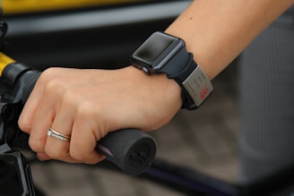 A hand gripping a bicycle handlebar is adorned with a smartwatch and a wristband. The smartwatch has a sleek, black band, and the wristband has a metallic tag. A simple ring with a small diamond is worn on the ring finger.