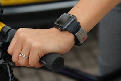 A hand gripping a bicycle handlebar is adorned with a smartwatch and a wristband. The smartwatch has a sleek, black band, and the wristband has a metallic tag. A simple ring with a small diamond is worn on the ring finger.