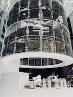 A modern coffee shop interior featuring a large, cylindrical glass display filled with coffee sacks labeled 'Arabica blend.' The lower section includes a sleek, white counter where baristas are preparing drinks, surrounded by minimalist coffee equipment and packaged coffee products.