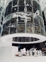 A modern coffee shop interior featuring a large, cylindrical glass display filled with coffee sacks labeled 'Arabica blend.' The lower section includes a sleek, white counter where baristas are preparing drinks, surrounded by minimalist coffee equipment and packaged coffee products.