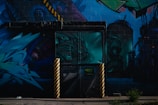 A vibrant urban mural featuring abstract art and graffiti. The mural includes various shades of blue and green, with details of a dragon-like creature, industrial elements, and geometric shapes. Yellow and black striped poles stand in front of a door painted with a complex design.