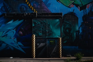 A vibrant urban mural featuring abstract art and graffiti. The mural includes various shades of blue and green, with details of a dragon-like creature, industrial elements, and geometric shapes. Yellow and black striped poles stand in front of a door painted with a complex design.