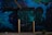 A vibrant urban mural featuring abstract art and graffiti. The mural includes various shades of blue and green, with details of a dragon-like creature, industrial elements, and geometric shapes. Yellow and black striped poles stand in front of a door painted with a complex design.