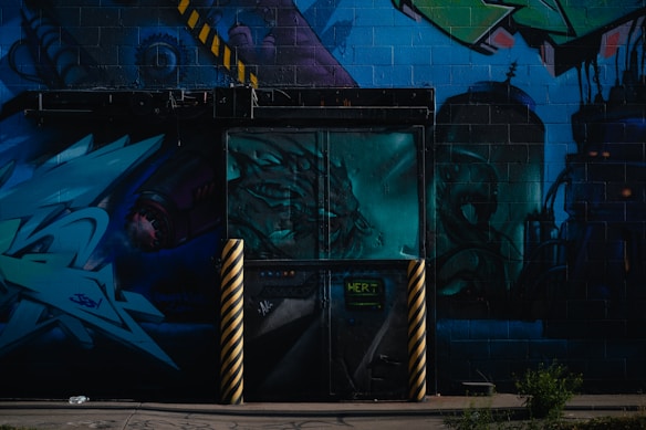 A vibrant urban mural featuring abstract art and graffiti. The mural includes various shades of blue and green, with details of a dragon-like creature, industrial elements, and geometric shapes. Yellow and black striped poles stand in front of a door painted with a complex design.