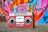 A vintage red boombox with cassette player and twin speakers is set against a vibrant graffiti-covered wall. The wall displays a mix of colors including pink, blue, and orange, enhancing the retro aesthetic.
