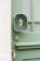 A close-up of a green door with a brass door handle, a keyhole, and an attached dark green plaque embossed with a golden symbol or letter.