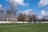 A soccer field with a green grass surface and a white goalpost situated to the left. The field is surrounded by tall, leafless trees, and a clear blue sky with scattered clouds creates a calm backdrop. Advertising banners are displayed along the fence bordering the field.