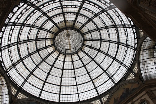A large circular glass dome with a geometric pattern consisting of multiple sections and metal framework. The dome is symmetrically designed with a central point from which the sections radiate outward. Surrounding the dome are ornate architectural details with decorative elements and artistic designs visible on the lower perimeter.