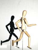 A wooden artist's mannequin is posed in a running motion, casting a shadow on a white background. The shadow mimics the mannequin's pose, adding a dynamic element to the scene. The mannequin's limbs are articulated, showcasing its flexibility.