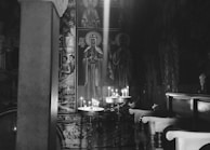 A dimly lit interior of a place of worship with religious icons on the walls. Lit candles are placed on a stand, casting a gentle glow. Wooden pews are positioned to the right, and the atmosphere is serene and contemplative.