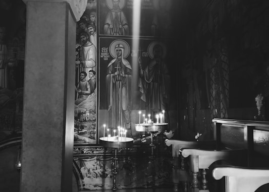 A dimly lit interior of a place of worship with religious icons on the walls. Lit candles are placed on a stand, casting a gentle glow. Wooden pews are positioned to the right, and the atmosphere is serene and contemplative.