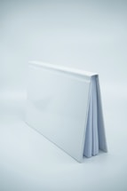 A white hardcover book is standing upright with its spine facing upward. The book's cover is glossy and reflects some light, and a few pages are partially visible.