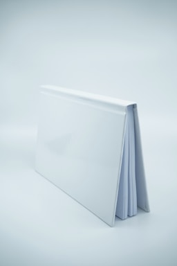 A white hardcover book is standing upright with its spine facing upward. The book's cover is glossy and reflects some light, and a few pages are partially visible.
