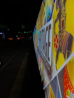 A colorful and vibrant street billboard with various artistic patterns and a stylized image of a figure in traditional attire. The words 'Heritage Liberty Plaza' are prominently displayed. The background is a nighttime urban street setting, and the sign is illuminated.
