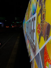 A colorful and vibrant street billboard with various artistic patterns and a stylized image of a figure in traditional attire. The words 'Heritage Liberty Plaza' are prominently displayed. The background is a nighttime urban street setting, and the sign is illuminated.