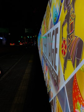 A colorful and vibrant street billboard with various artistic patterns and a stylized image of a figure in traditional attire. The words 'Heritage Liberty Plaza' are prominently displayed. The background is a nighttime urban street setting, and the sign is illuminated.