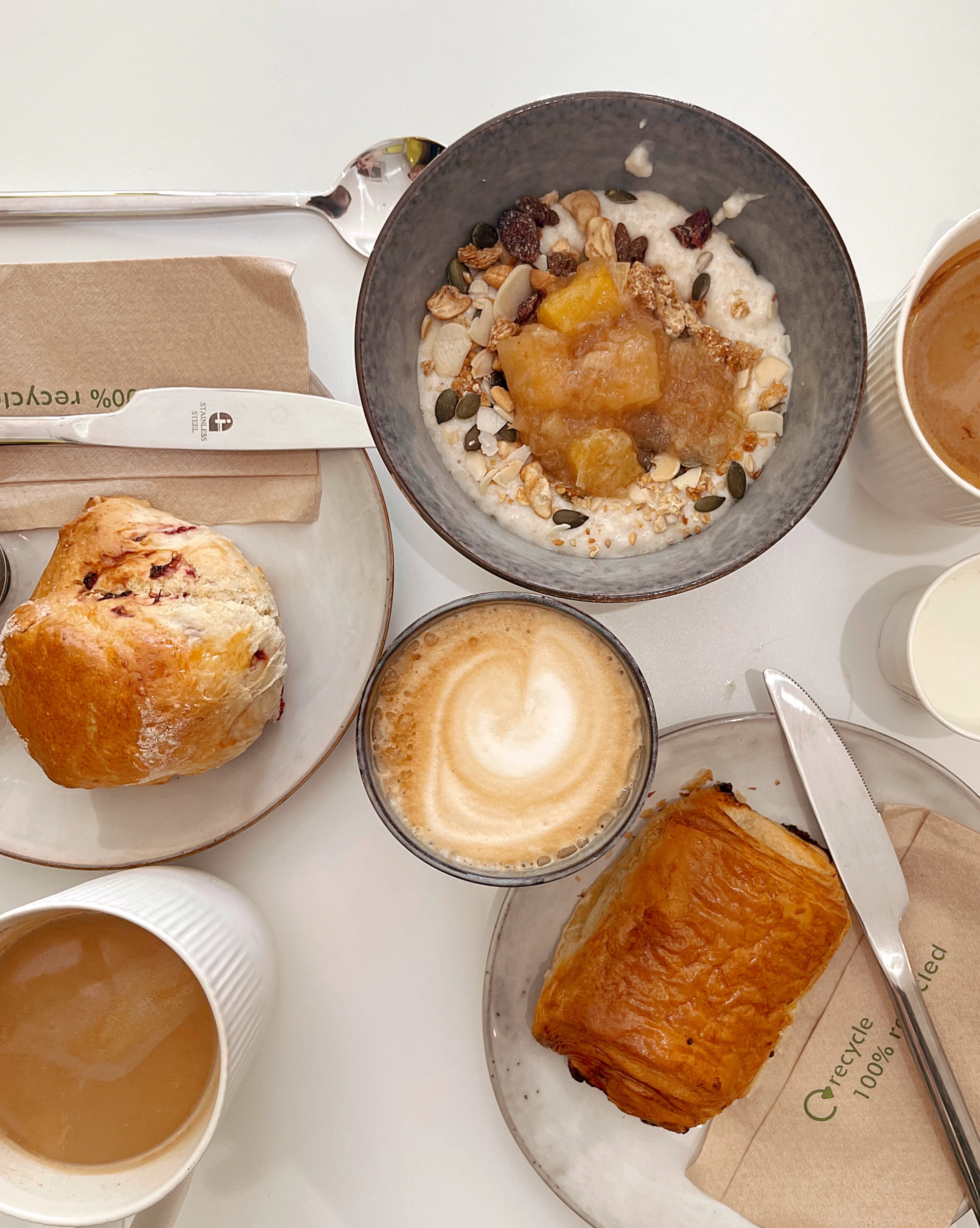 A Belfast cafe table from above: a fruit scone, a pain au chocolat, a bowl of porridge with poached fruit and seeds, and a latte with rosetta art