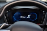 A car dashboard display featuring the BMW i logo prominently in the center with circular graphic elements in blue. The steering wheel partially frames the display, and the interior appears modern with a digital interface.