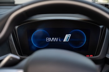 A car dashboard display featuring the BMW i logo prominently in the center with circular graphic elements in blue. The steering wheel partially frames the display, and the interior appears modern with a digital interface.