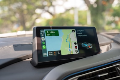 A car dashboard featuring a digital display screen. The screen shows a navigation map with route details, including the distance to the destination and estimated time of arrival. It also indicates the car's battery charge status and energy flow. The interior of the car is modern with a grey color scheme.