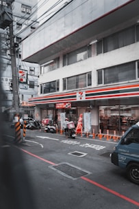 A convenience store with a recognizable sign is situated in an urban street. Delivery scooters and a person wearing a helmet are visible in front, along with a parked vehicle. Orange traffic cones line the sidewalk, and various signs and advertisements are present on the store’s facade and windows.