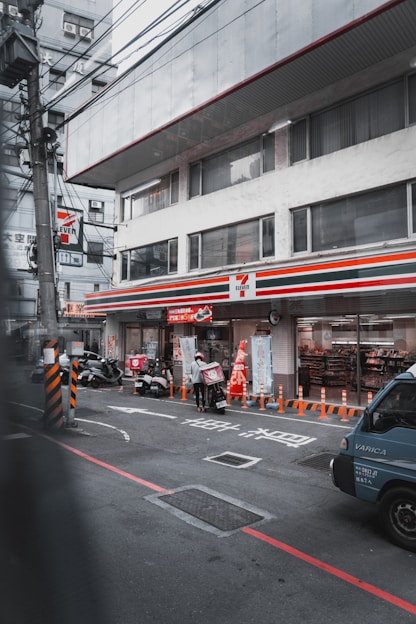 A convenience store with a recognizable sign is situated in an urban street. Delivery scooters and a person wearing a helmet are visible in front, along with a parked vehicle. Orange traffic cones line the sidewalk, and various signs and advertisements are present on the store’s facade and windows.