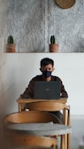 A person wearing a black mask is working on a laptop at a wooden table in a minimalist setting with a textured wall and two small potted cacti on a shelf behind.