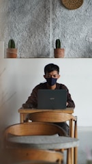 A person wearing a black mask is working on a laptop at a wooden table in a minimalist setting with a textured wall and two small potted cacti on a shelf behind.