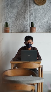 A person wearing a black mask is working on a laptop at a wooden table in a minimalist setting with a textured wall and two small potted cacti on a shelf behind.