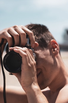 Close-up of a person holding a camera to their eye, focusing on taking a photograph. The background is blurred, suggesting an outdoor setting with bright lighting.