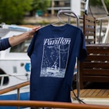 A hand is holding a navy blue T-shirt with a design featuring a boat and navigation lines, along with the word 'Pavillon.' The background includes a boat deck with wooden furniture and a partially visible white and blue boat.
